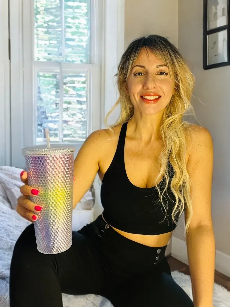 Lena à la maison, brassière noire, un grand gobelet irisé à la main, sourire