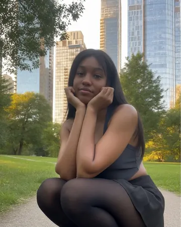 Maya crouching in a park below tall buildings, tights and ankle boots