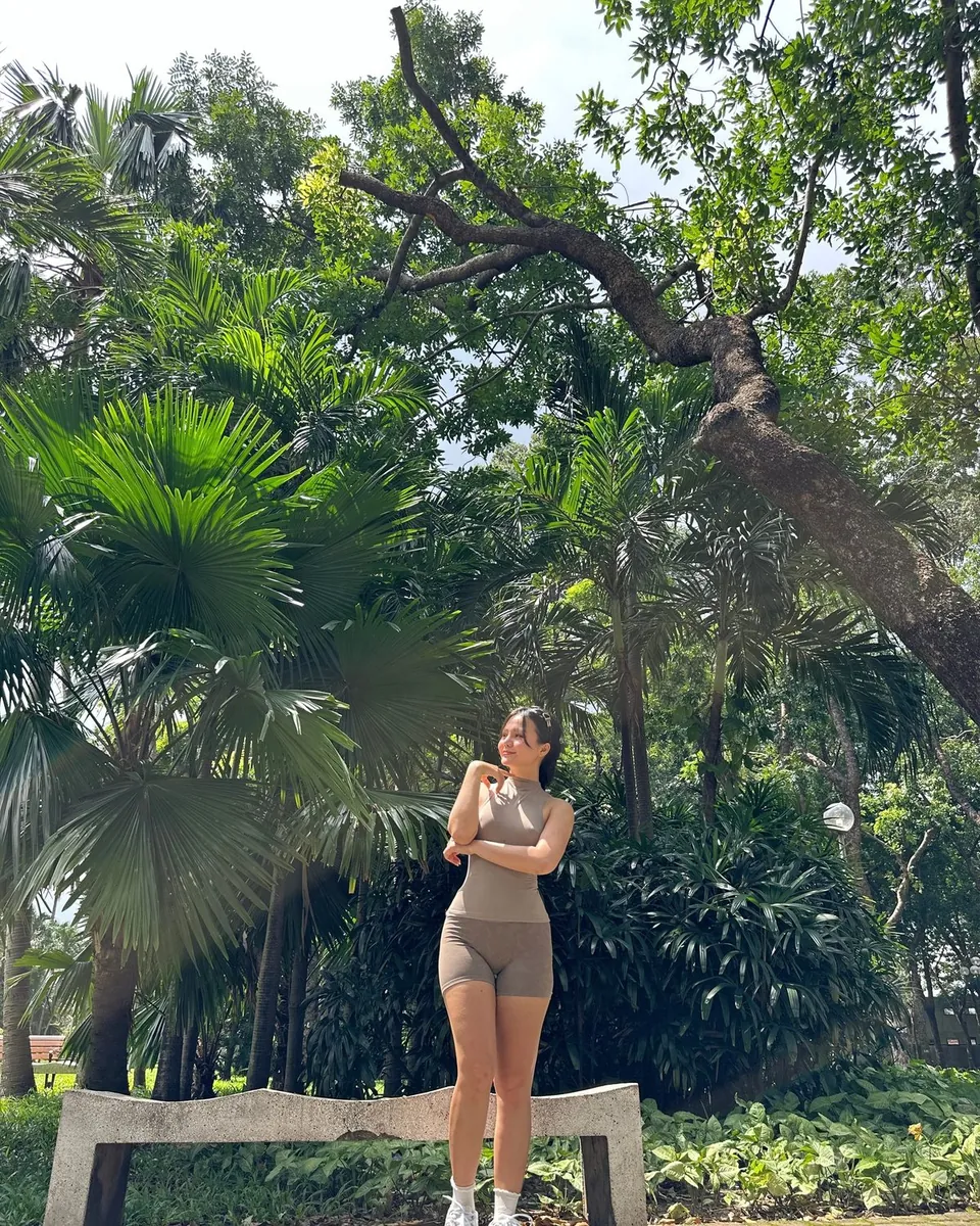 Mia debout près d'un banc de pierre dans un parc aux arbres tropicaux, tenue de sport beige