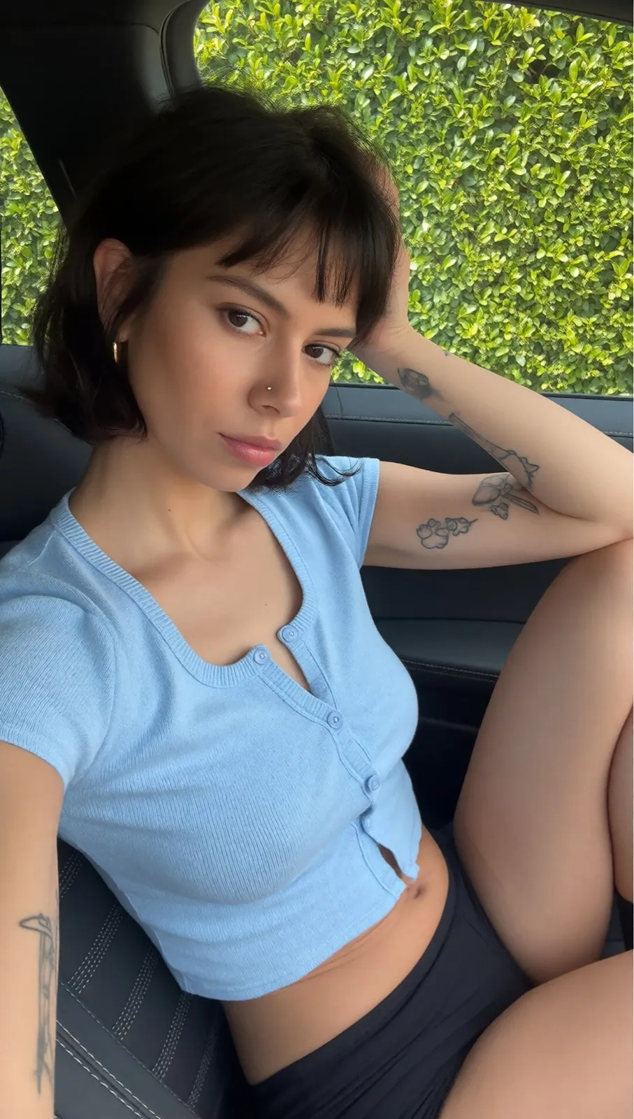 Romy in a car, short brown bob with a fringe, light blue top, fine tattoos on her arm