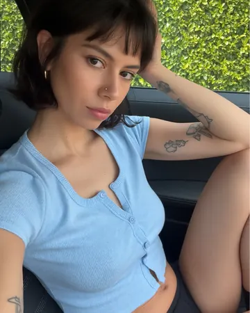 Romy in a car, short brown bob with a fringe, light blue top, fine tattoos on her arm