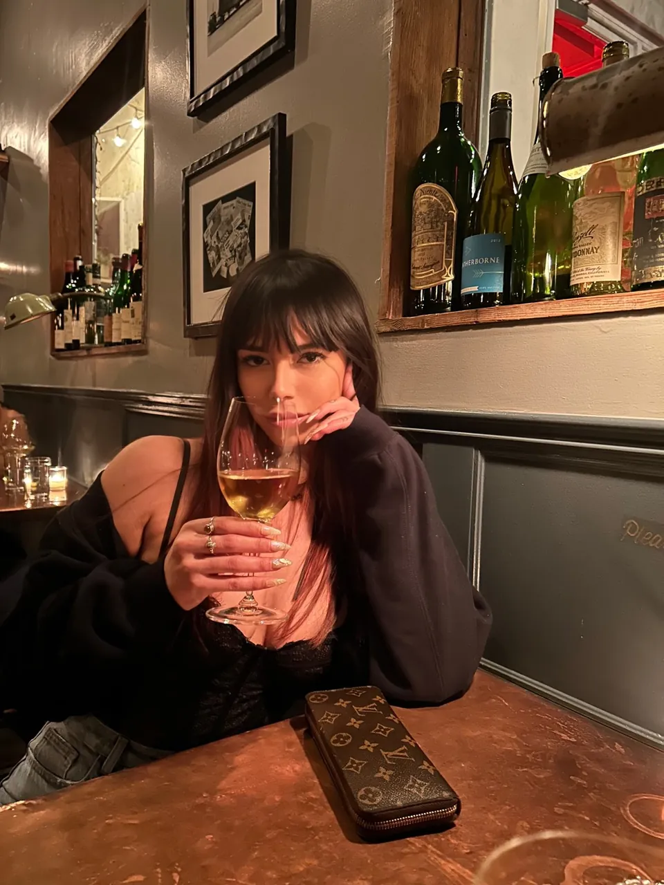 Romy assise à une table en bois dans un bar, un verre de vin à la main, pull noir