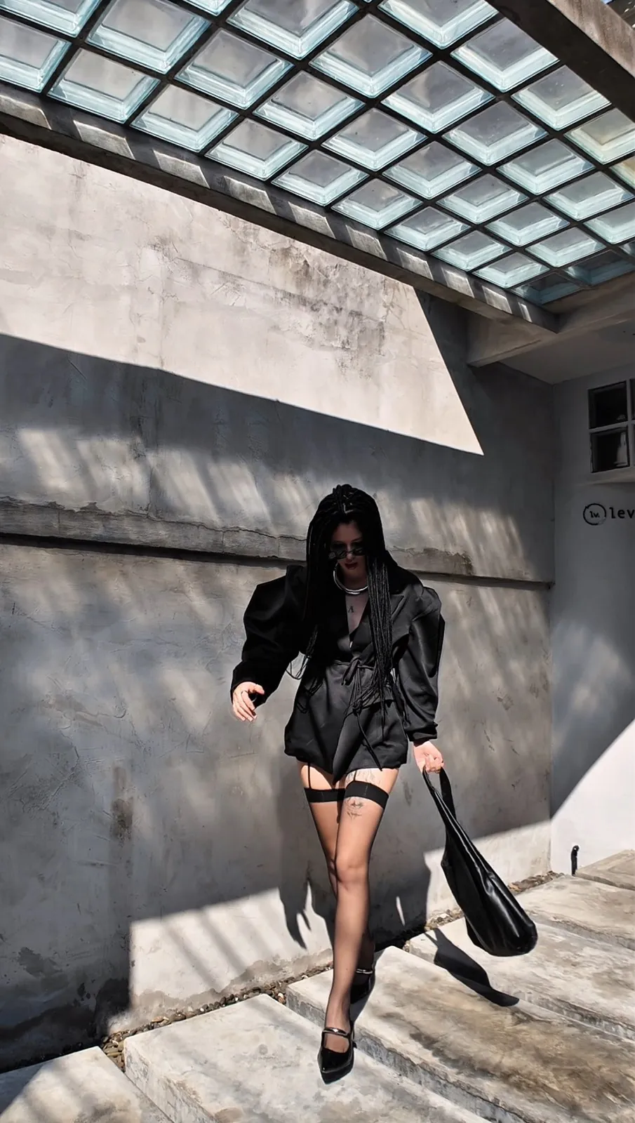 Vera devant un mur de béton gris, veste noire et lunettes de soleil